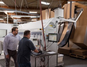Two men, one standing and one seated, are interacting at a computer terminal in a manufacturing plant with large machinery in the background.