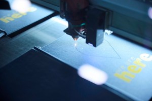 A close-up shot of a laser engraving machine in operation. A tiny, bright dot of light is visible as a laser head moves across a dark surface, leaving a faint line behind it. Another piece of material with the yellow text "here" is out of focus in the foreground, suggesting a precise, automated process.| Platinum Equity