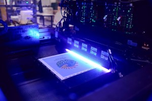A detailed shot of a printing press in operation. A piece of paper with a colorful design is visible on a conveyor belt under a bright, glowing blue light. The machine's internal components, including circuit boards with green lights and various wires, are in the background. The overall scene is dark with the machinery illuminated by the blue and green lights. | Platinum Equity