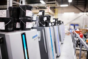 A detailed shot of printing machinery in a factory. Several black buckets of ink are stacked on top of a gray machine labeled "HEIDELBERG." The machine has a blue vertical light strip and a digital display. Other similar machines are lined up in the background, creating a sense of a large-scale production line. | Platinum Equity