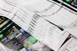 A close-up shot of stacks of printed yearbook pages. The pages are fanned out, revealing various layouts with photos of students playing sports and other activities. Barcodes and timestamps are visible on the edges of some pages, indicating they are fresh off the printing press. The pages are a mix of full-color and black-and-white. | Platinum Equity