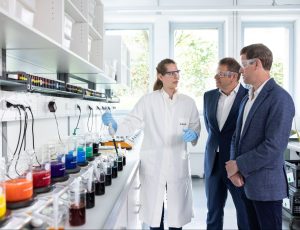 female scientist talking to two men in suites while pointing to lab equipment