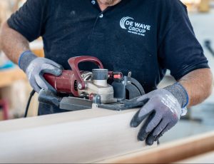 A person wearing gloves is using a red power tool to sand or smooth a piece of light-colored wood, while wearing a black shirt with the "DE WAVE GROUP" logo. | Platinum Equity