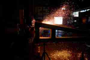 A welder wearing a helmet and protective gear is working in a dark, industrial setting. Bright orange and yellow sparks fly from the metal beam being worked on, illuminating the surrounding area. | Platinum Equity