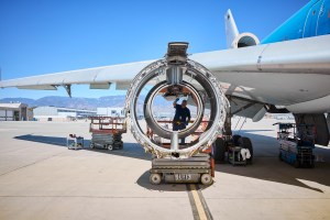 A full shot of an airplane engine being worked on by a technician. The engine's outer casing is removed, revealing the inner workings. The technician is standing on a scissor lift platform inside the engine's frame. The main body and wing of a large passenger plane are visible in the background, parked on an outdoor tarmac under a clear blue sky. | Platinum Equity