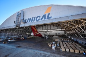 A wide-angle view of a large aircraft hangar. A plane with a red tail is parked inside, surrounded by stacked boxes and other equipment. The word "UNICAL" and a stylized orange logo are prominently displayed on the exterior of the hangar. | Platinum Equity