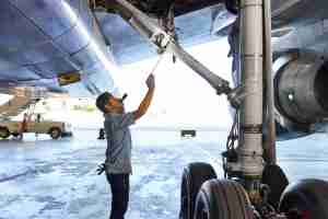 A male aircraft technician is working under the wing of a large airplane. He is holding a tool and looking up at the landing gear mechanism. The landing gear tires and part of an engine are visible, and the scene is set inside a hangar with a concrete floor. | Platinum Equity
