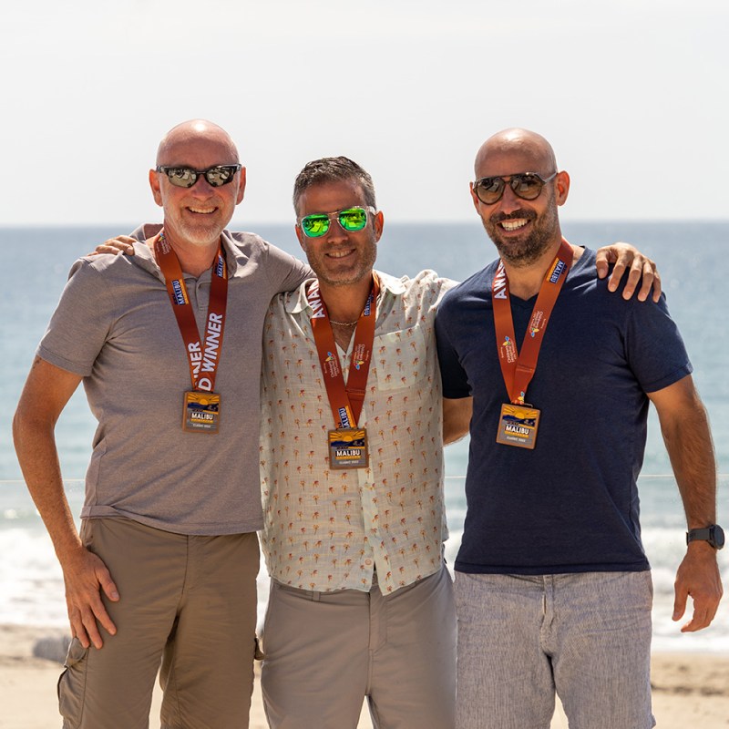 Three individuals wearing medals, standing in a beach-like setting with a summery ambiance