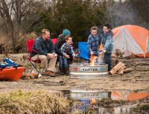 A family is camping outdoors, roasting marshmallows over a Tarter-branded fire ring, with tents and a kayak nearby | Platinum Equity