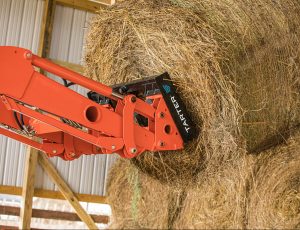A close-up view of a bright orange tractor attachment, branded with the Tarter logo, lifting a large round bale of hay inside a barn. The hay spear is securely embedded in the bale, with wooden beams and a corrugated metal roof visible in the background. | Platinum Equity
