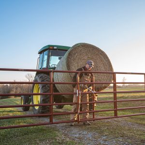 Photo depicting a farmer opening a red gate while a tractor lifts a cylindrical bundle of grass. | Platinum Equity