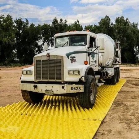 A white cement mixer truck driving over a yellow, textured plastic spill containment mat in a dusty area with trees in the background.