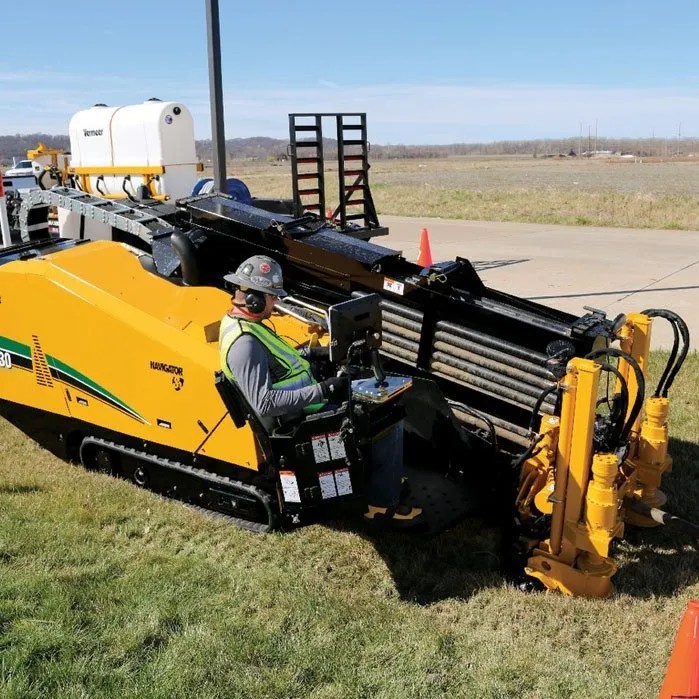 A worker in reflective gear operates a large, yellow trencher machine on a grassy terrain under a clear sky.