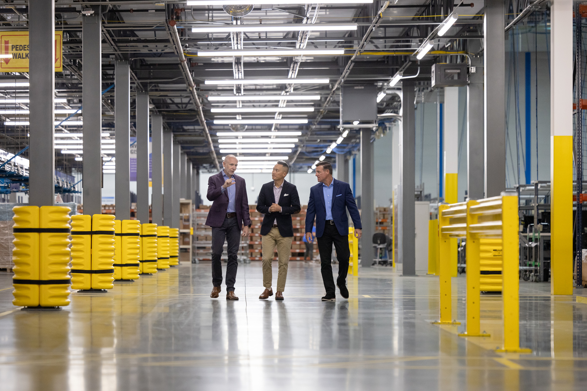 Three business professionals, two men and one woman, walk through a large industrial warehouse with yellow containers and structural columns, engaged in a conversation.