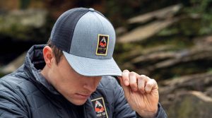 A person in a quilted jacket is adjusting the brim of a gray-and-black mesh cap with a "High Top State Park Staff" patch on the front. | Platinum Equity