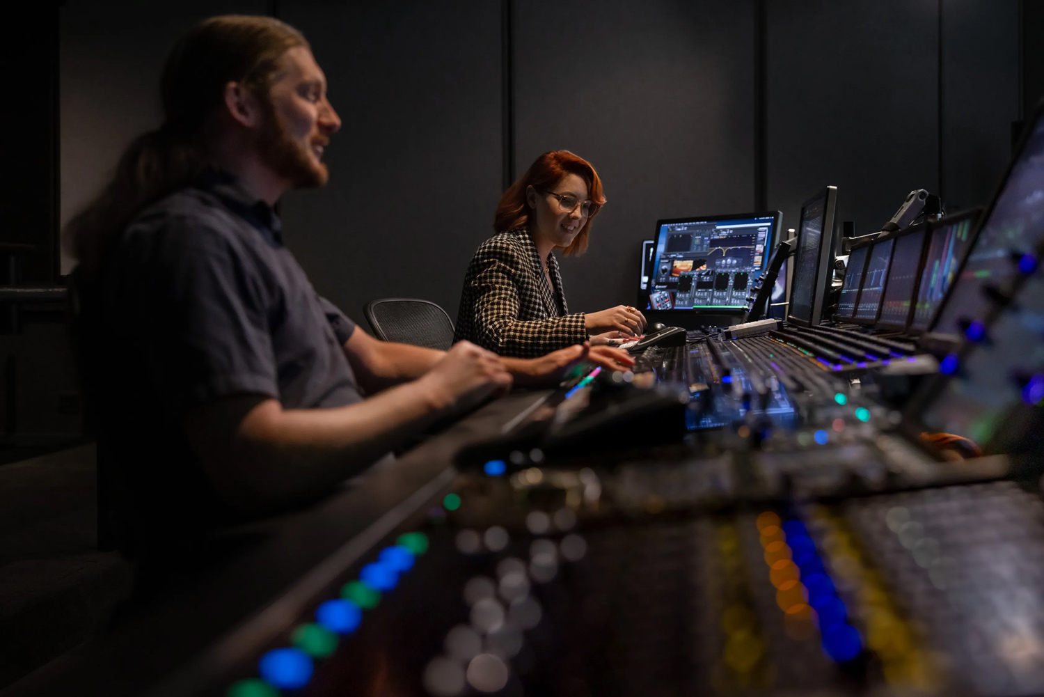 The image shows a man and a woman working on a DJ set in a recording studio. They are using a mixing console and other audio equipment to create music. The scene includes a computer and various electronic devices commonly used by DJs.