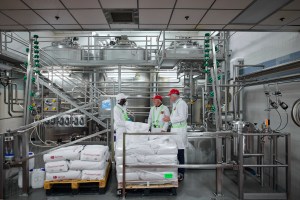Three individuals in lab coats and hard hats are standing and talking in a food processing facility. They are gathered in front of large stainless steel tanks and complex machinery, with pipes and railings filling the background. | Platinum Equity
