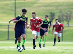 Two soccer players in opposing team uniforms race toward the ball, with teammates chasing closely behind on a grassy field. | Platinum Equity