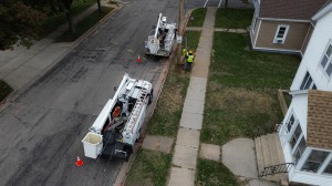 An aerial shot of a residential street with two utility trucks. The trucks are parked on the road, and a crew of people in high-visibility vests and hard hats are working on a utility pole. The scene includes sidewalks, a grassy lawn, and parts of two houses on the right. | Platinum Equity