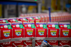 A panoramic shot of a row of red and white cartons of "Horizon Organic Half and Half" milk. Each carton features the brand's logo and a drawing of a cow jumping over the moon. The white caps and product information are clearly visible, and the cartons are arranged neatly side-by-side. | Platinum Equity