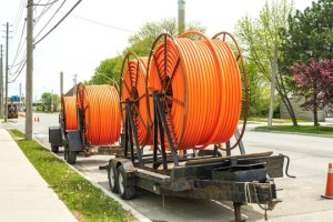 A series of three large trailers, each carrying a massive spool of orange conduit piping, are parked along a street. The spools are connected to a long pipeline that runs down the street, suggesting construction or utility work. The setting is a suburban street with sidewalks, grass, and trees. | Platinum Equity