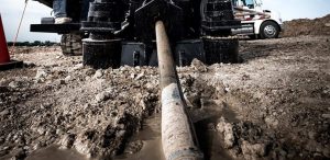 A low-angle shot of a drilling rig at a construction site. The drill pipe is submerged in a muddy, wet trench in the foreground. The rig's machinery and the tires of a large truck are visible in the background. The ground is a mix of mud, dirt, and water. | Platinum Equity