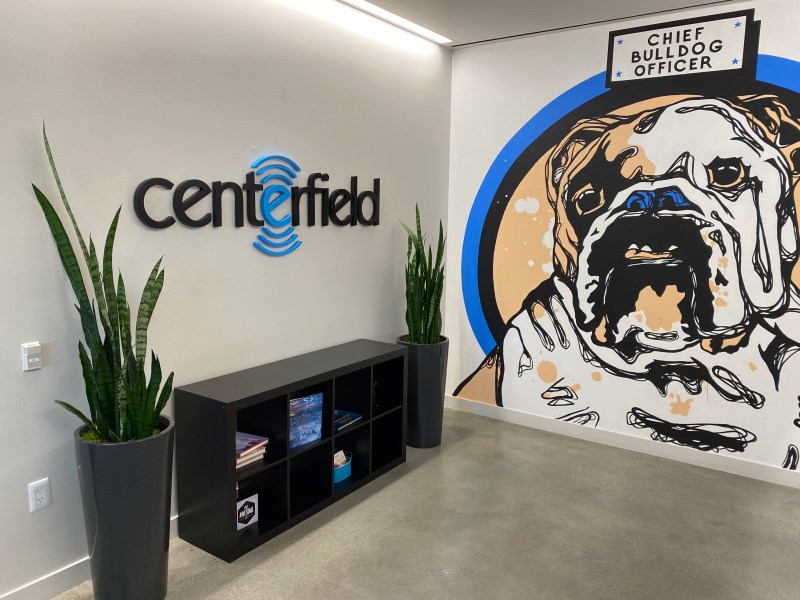 A corner of a modern office lobby. The "Centerfield" logo is mounted on a light gray wall, with the stylized logo in bright blue neon. To the right, a large, stylized black and white mural of a bulldog is painted on the wall, with the text "CHIEF BULLDOG OFFICER" above it. A black shelf unit is placed between two tall snake plants in planters. | Platinum Equity