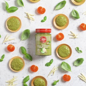 A top-down shot of a jar of pesto sauce without garlic, surrounded by ingredients and small dishes of the sauce on a white textured surface. The jar is the central focus, with cherry tomatoes, fresh basil leaves, and pieces of pasta scattered around it. Small wooden bowls hold dollops of the green pesto, creating a stylized and appetizing arrangement. | Platinum Equity