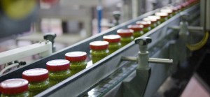 A long, diagonal shot of a conveyor belt in a factory. Rows of glass jars filled with pesto sauce. Each jar is sealed with a red lid featuring a white logo. | Platinum Equity