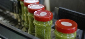 close-up shot of a factory conveyor belt. Rows of glass jars filled with green pesto sauce are moving from the top left to the bottom right of the frame. Each jar is sealed with a red lid featuring a white brand logo. | Platinum Equity