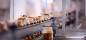 A low-angle shot of a conveyor belt in a factory, showing rows of glass jars filled with tomato-based sauce and are moving along a production line. | Platinum Equity