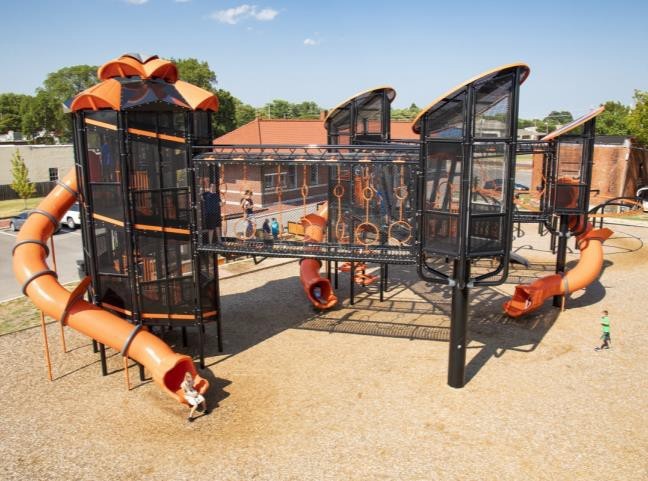 Children playing on a modern outdoor playground with climbing structures, swings, and colorful equipment surrounded by trees and open space. | Platinum Equity