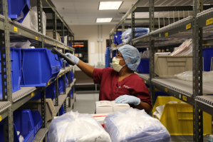 Worker wearing scrubs, a mask, gloves, and a hair cover uses a handheld scanner to check items on shelves filled with blue storage bins in a supply room. | Platinum Equity