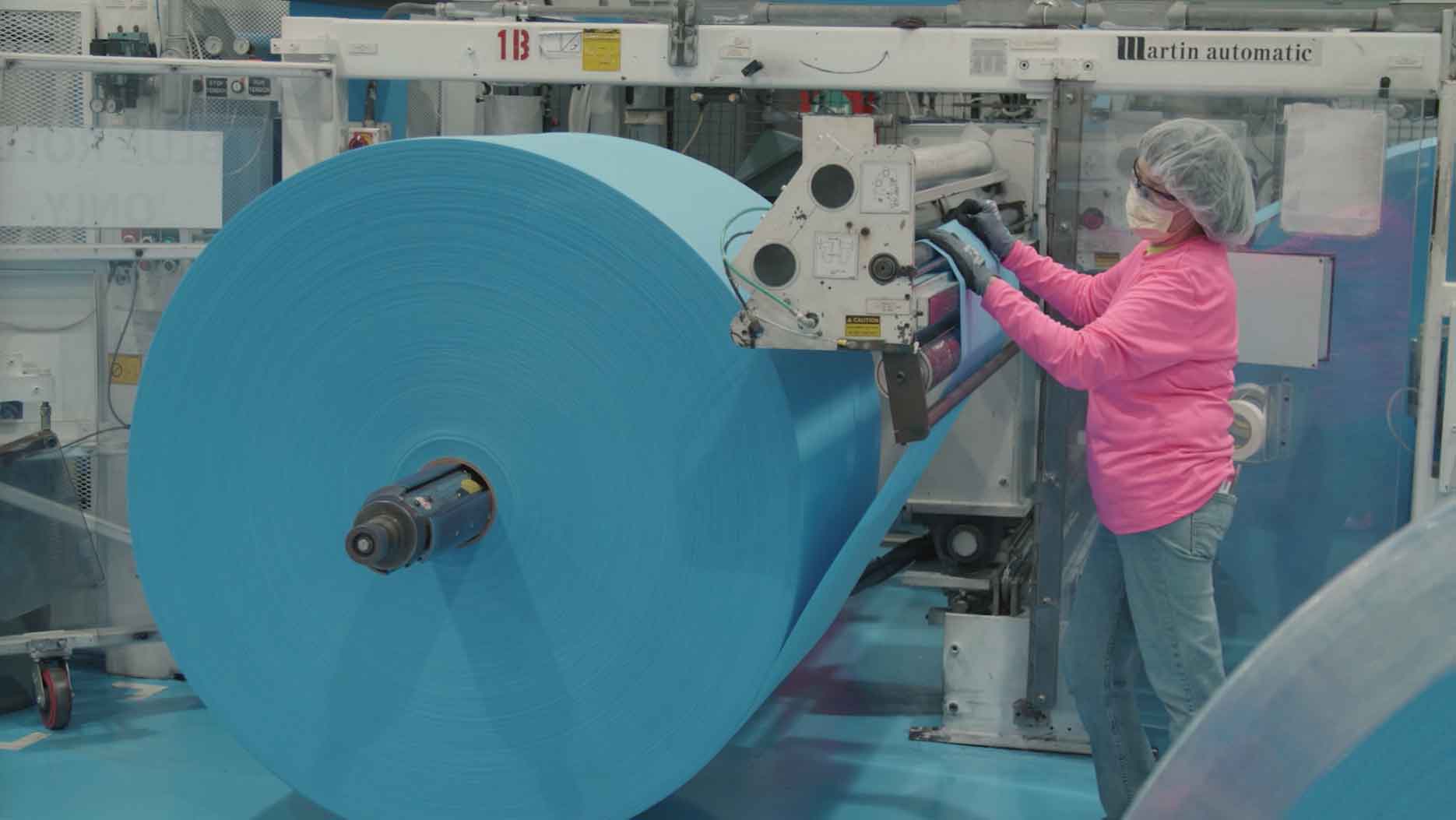 Worker wearing protective gear operates industrial equipment while guiding a large blue roll of material through a manufacturing machine on a factory floor | Platinum Equity
