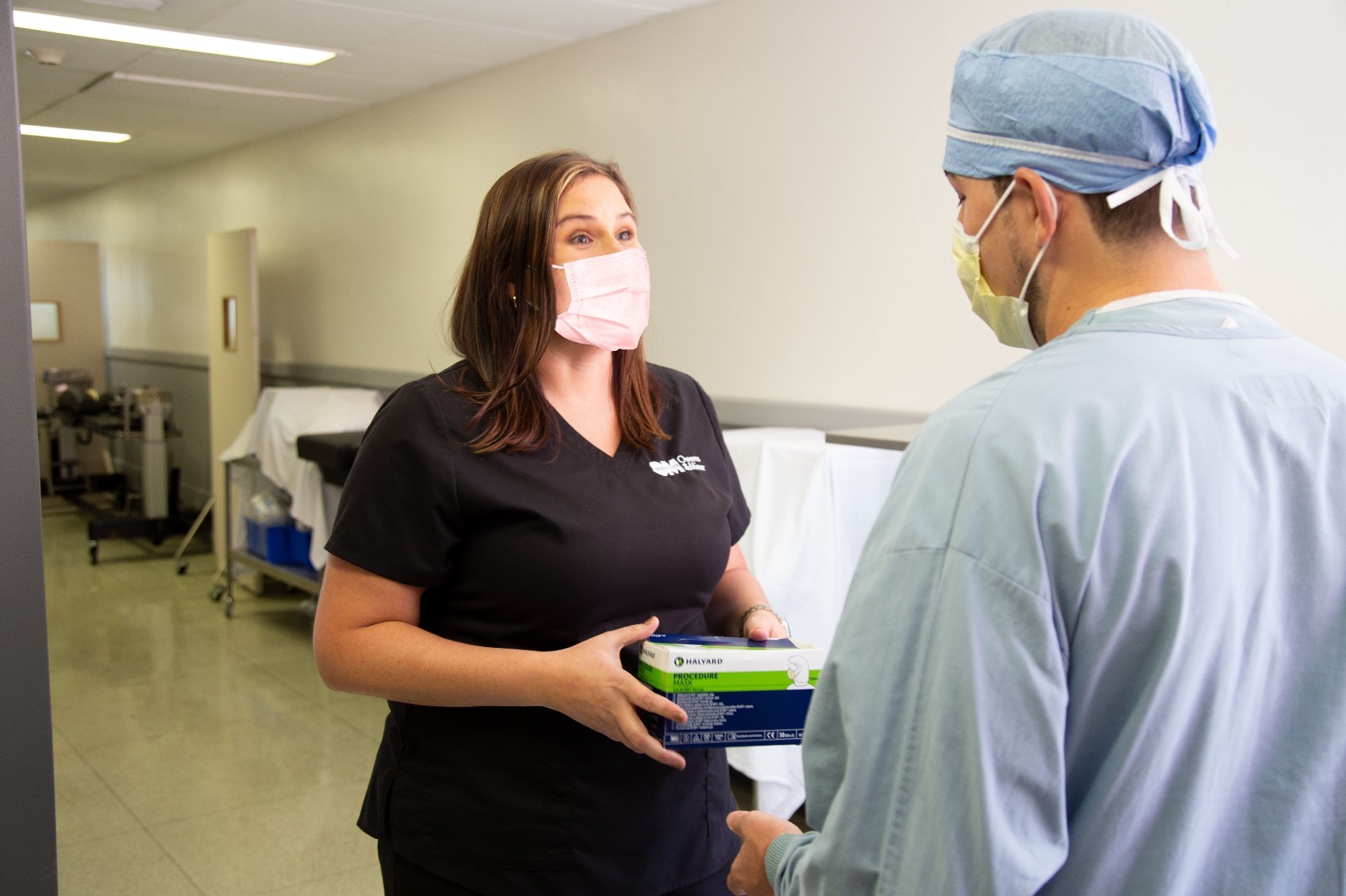 Healthcare worker wearing a face mask hands a box of medical gloves to a clinician in scrubs and a surgical cap while standing in a hospital hallway | Platinum Equity