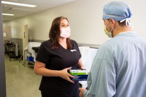 Healthcare worker wearing a face mask hands a box of medical gloves to a clinician in scrubs and a surgical cap while standing in a hospital hallway | Platinum Equity