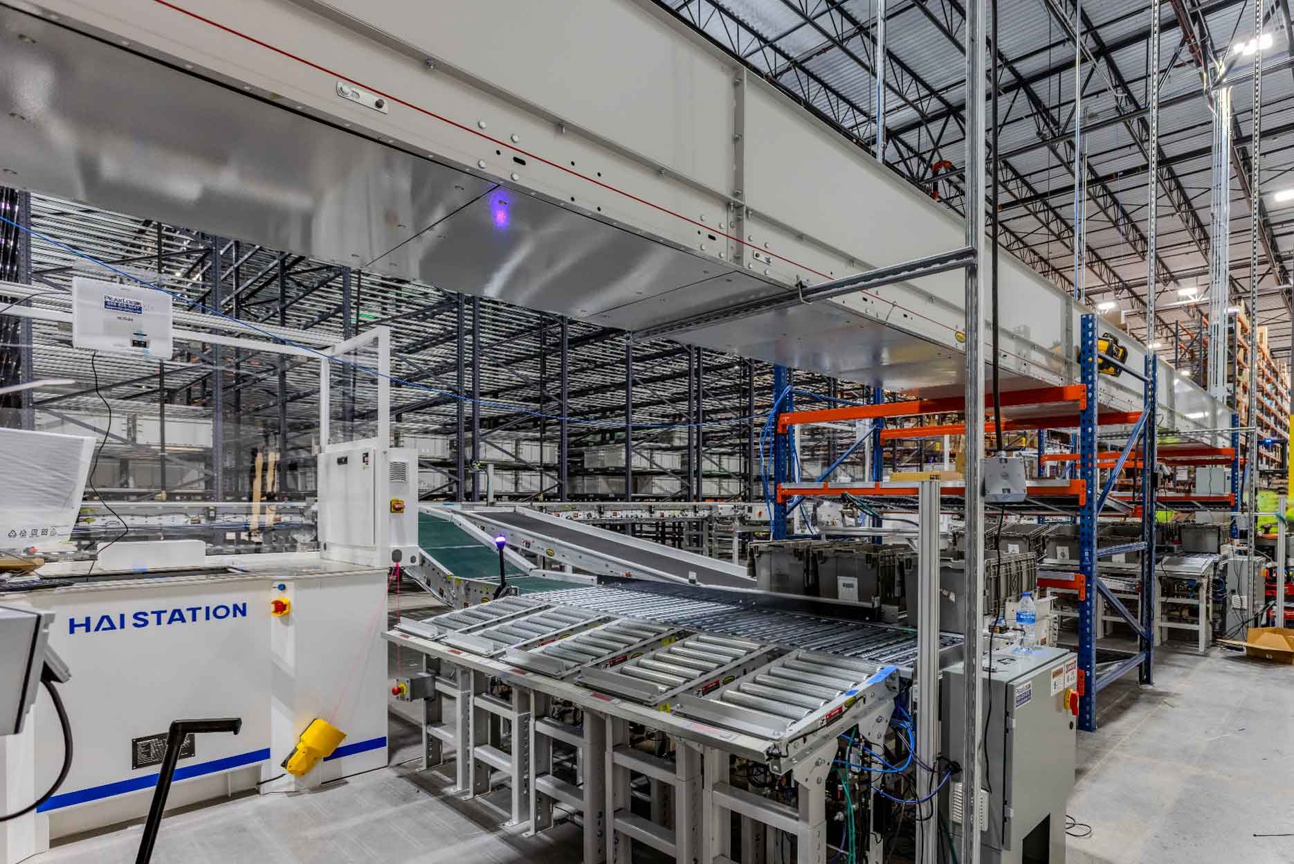 Automated conveyor and sorting system inside a large warehouse, showing metal rollers, overhead equipment, and tall storage racks arranged across the facility | Platinum Equity