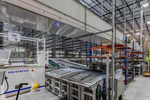 Automated conveyor and sorting system inside a large warehouse, showing metal rollers, overhead equipment, and tall storage racks arranged across the facility | Platinum Equity