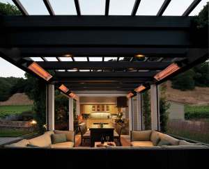 Covered outdoor patio with black pergola, glowing overhead heaters, cushioned seating, and a dining table leading to an illuminated outdoor kitchen at dusk, surrounded by landscaped hills and greenery. | Platinum Equity
