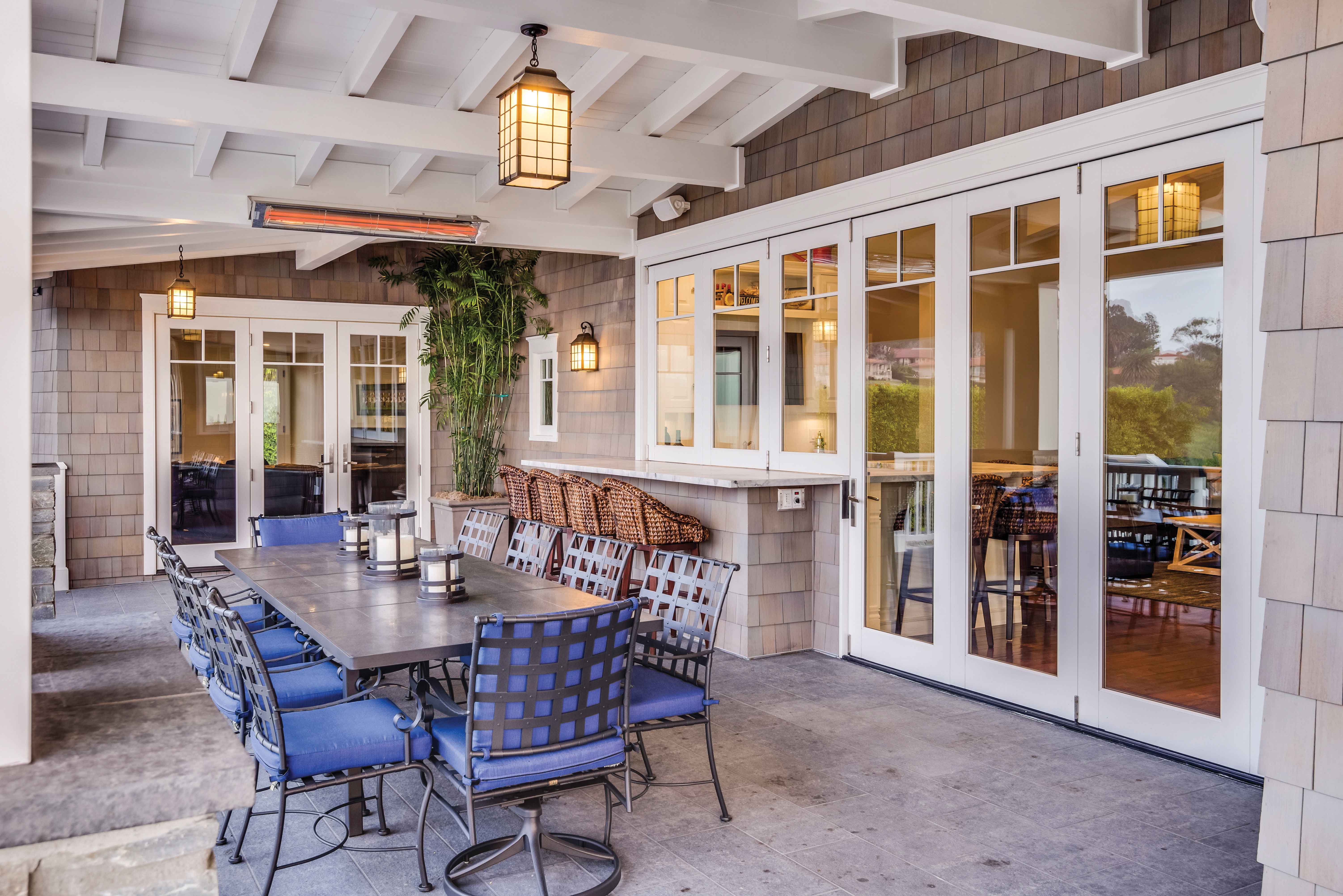 Covered outdoor dining patio with a long table, blue cushioned chairs, hanging lantern lights, white-framed glass doors, and a mounted infrared heater beneath a white-beamed ceiling. | Platinum Equity