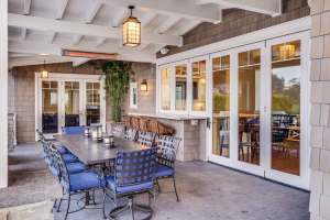 Covered outdoor dining patio with a long table, blue cushioned chairs, hanging lantern lights, white-framed glass doors, and a mounted infrared heater beneath a white-beamed ceiling. | Platinum Equity