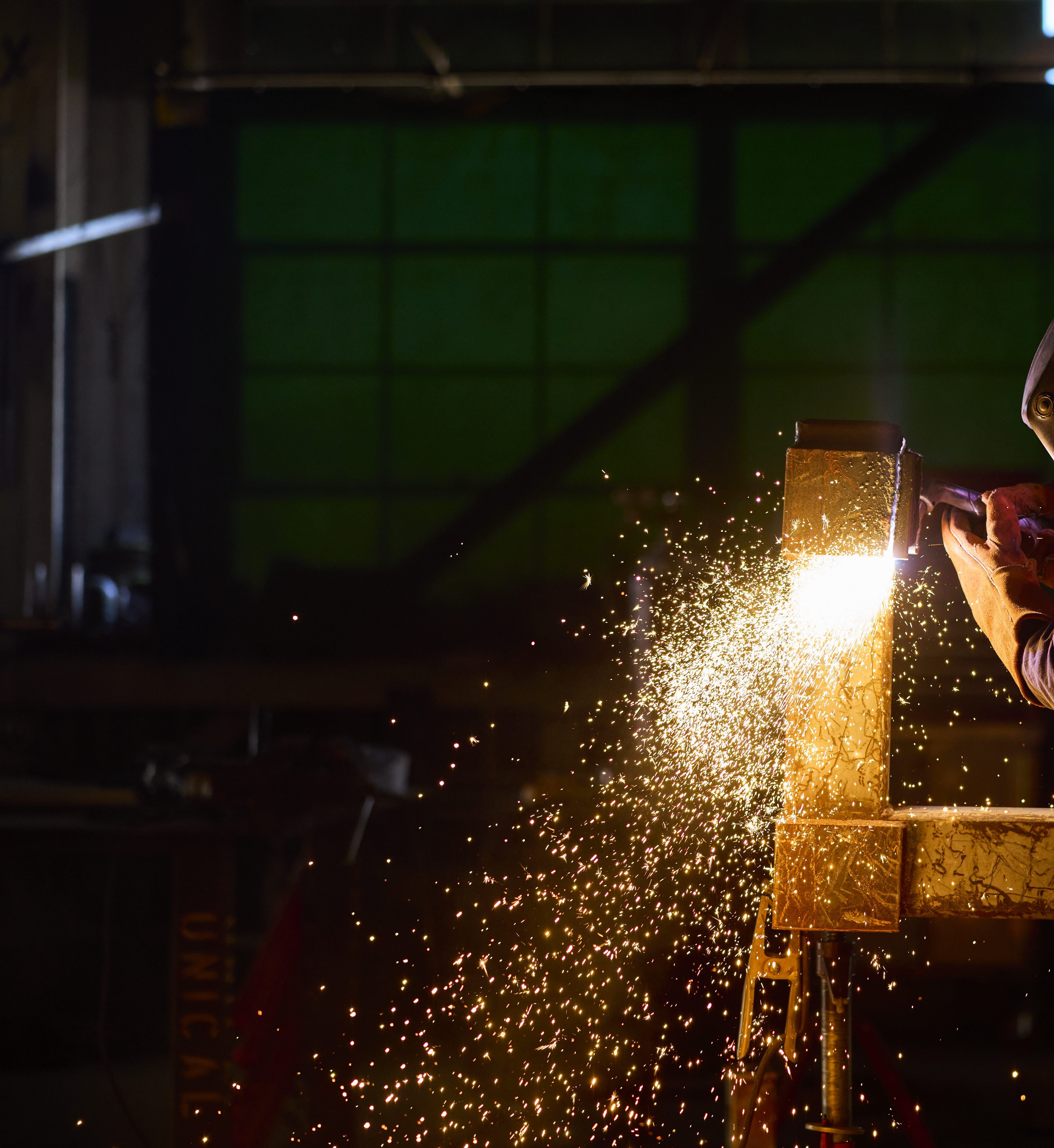 A worker wearing a welding helmet uses welding equipment on a large metal beam, with bright sparks illuminating an industrial workshop. | Platinum Equity
