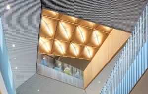 An interior atrium features geometric wood ceiling panels with integrated lighting, metal railings, and people gathered on an upper-level walkway. | Platinum Equity